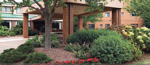 Covered entrance of a brick senior living building with a canopy, trees, shrubs, and flower beds.
