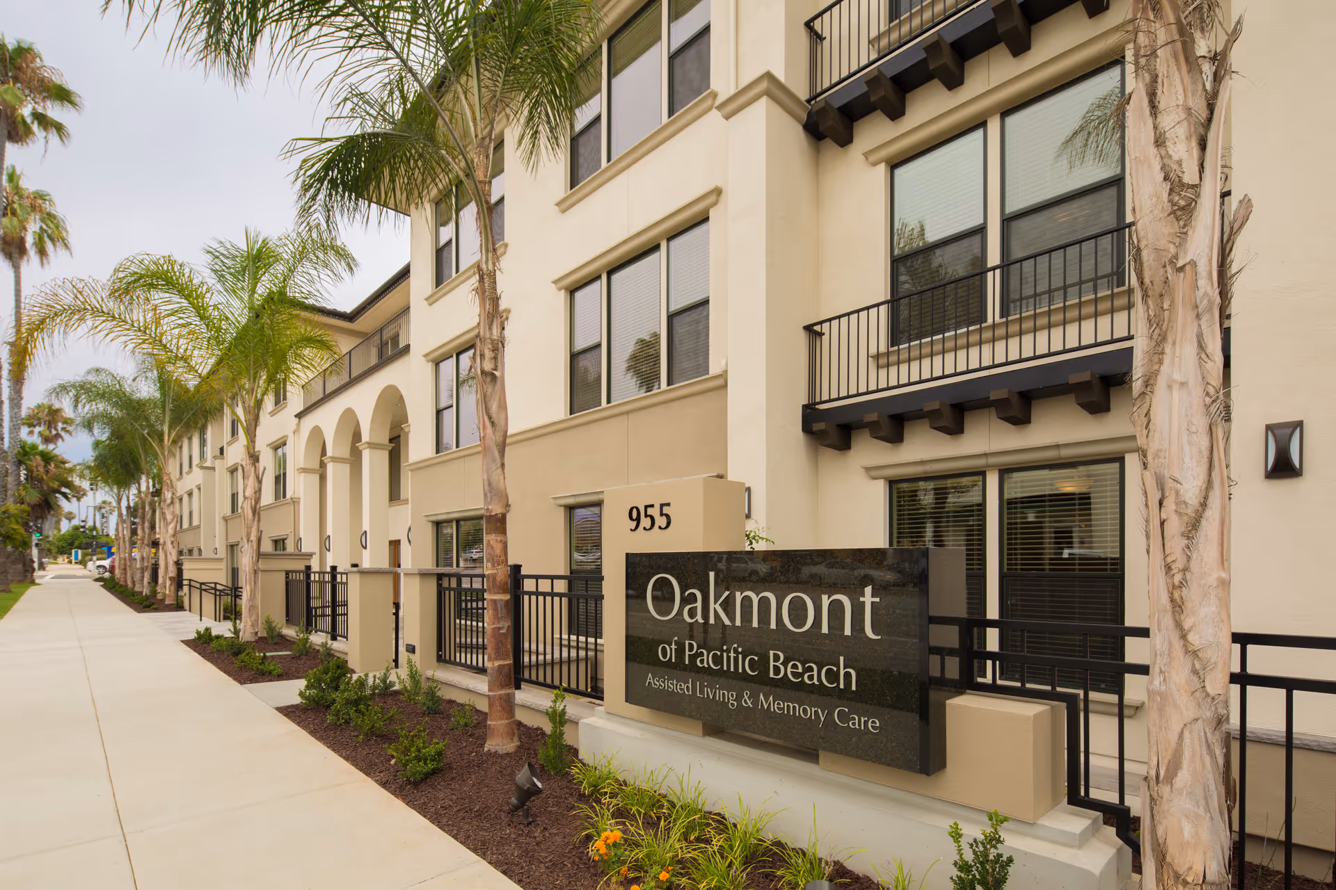 Exterior view of Oakmont of Pacific Beach, a beige multi-story assisted living and memory care facility with palm trees lining the sidewalk and a large sign displaying the facility name and address number 955.