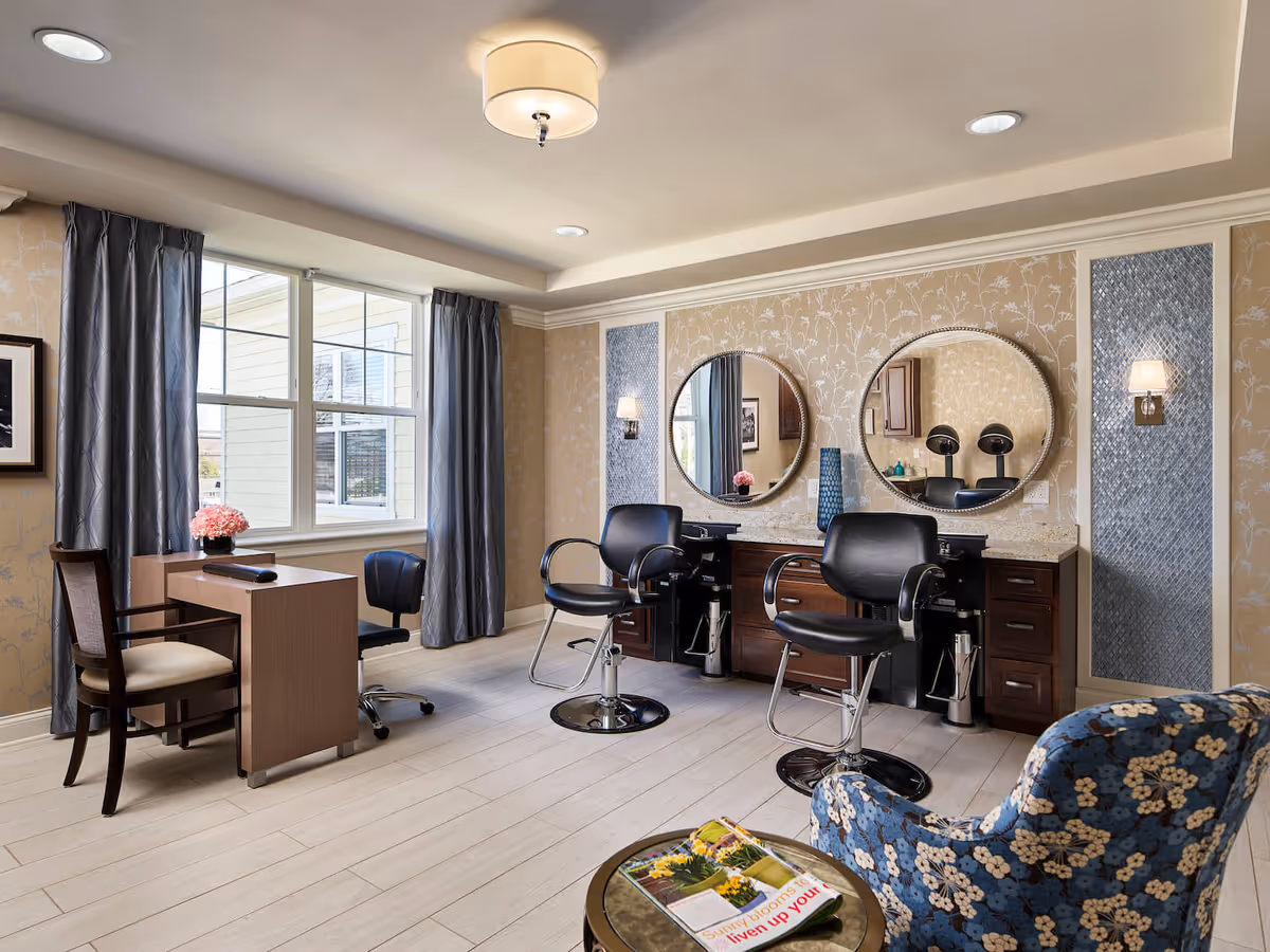 A well-lit salon area in an assisted living facility featuring two black salon chairs in front of a counter with two round mirrors mounted on a beige floral wallpapered wall. There is a window with gray curtains letting in natural light, a small table with a flower vase, and a blue floral upholstered chair next to a small round table with magazines.