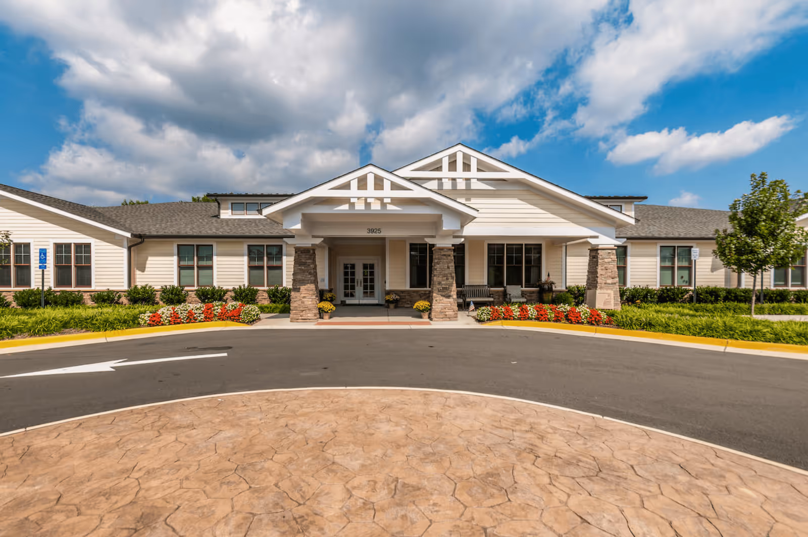 Front exterior view of Chantilly Heights Memory Care facility with a covered entrance supported by stone pillars, surrounded by well-maintained landscaping and colorful flowers under a partly cloudy sky.