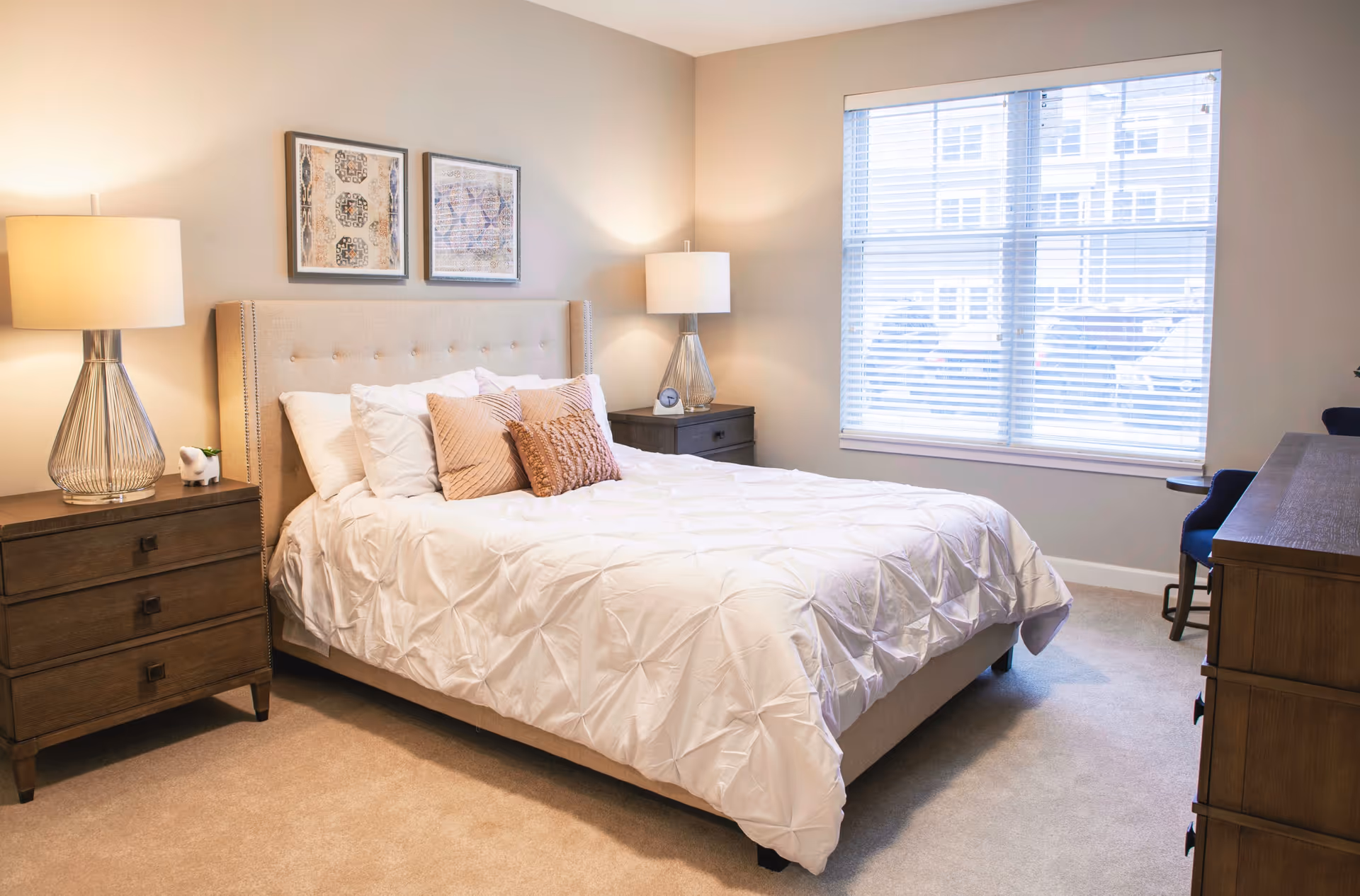 A bright bedroom with a beige upholstered bed featuring white bedding and decorative pillows. There are two wooden nightstands with lamps on either side of the bed. Two framed artworks hang above the headboard. A large window with blinds allows natural light to fill the room. A wooden dresser and a blue chair are visible on the right side.