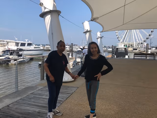 Two women holding hands and standing on a waterfront boardwalk with boats docked in the marina and a large Ferris wheel in the background under a white canopy structure.