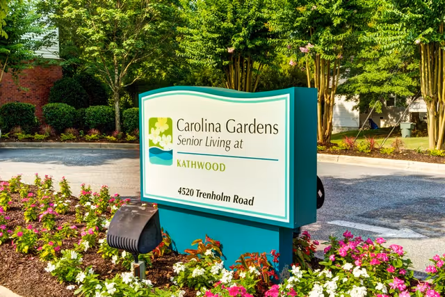 Outdoor sign for Carolina Gardens Senior Living at Kathwood located at 4520 Trenholm Road, surrounded by landscaped flowers and greenery with trees and a parking area in the background.