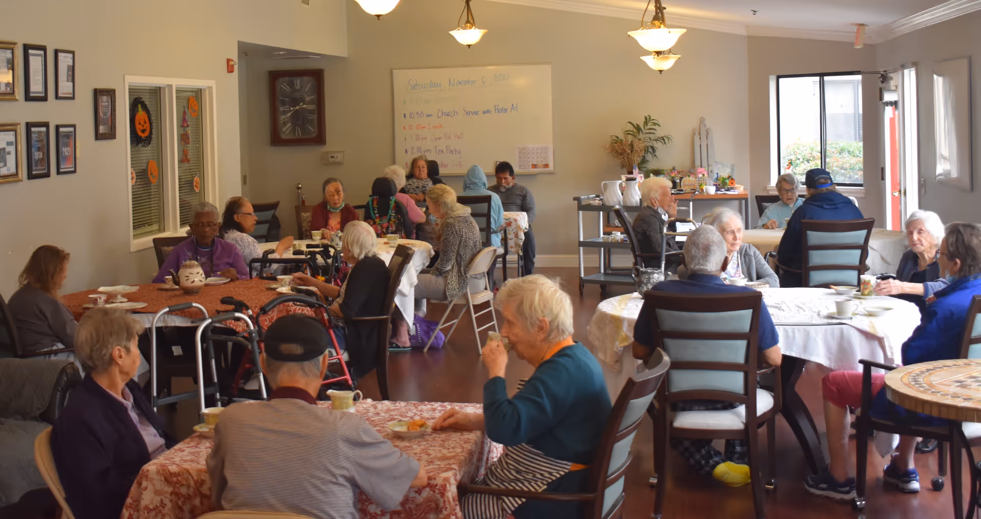 A group of elderly people sitting at multiple round tables in a well-lit dining room, engaging in conversation and having tea or coffee. The room has light-colored walls, a large clock, a whiteboard with a schedule, and some decorations on the walls and tables.