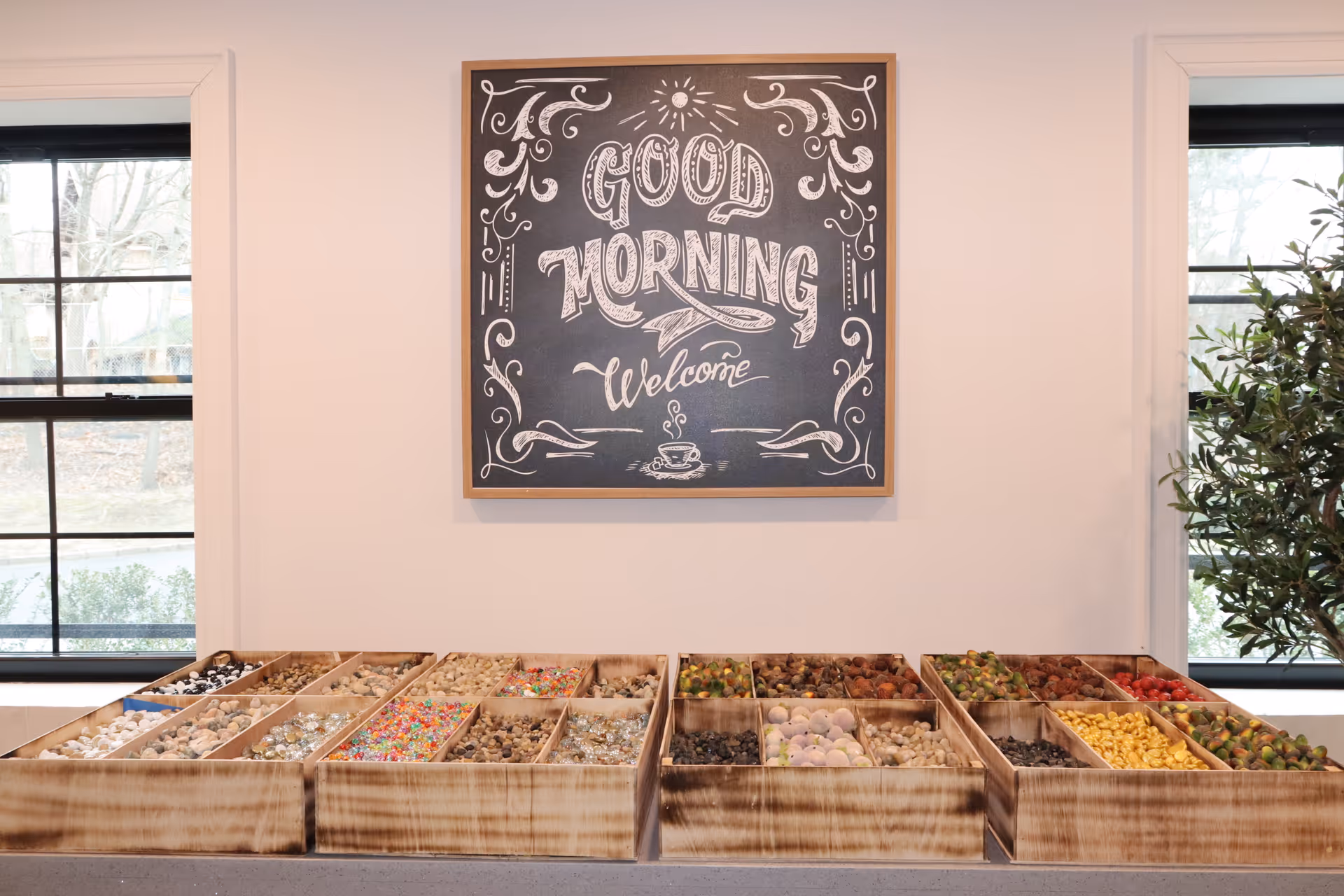 A wooden display box with multiple compartments filled with various colorful decorative stones and beads is placed on a table. Above it, a framed chalkboard-style sign reads 'Good Morning Welcome' with decorative flourishes. Two windows with black frames and a green plant are visible on either side of the sign.