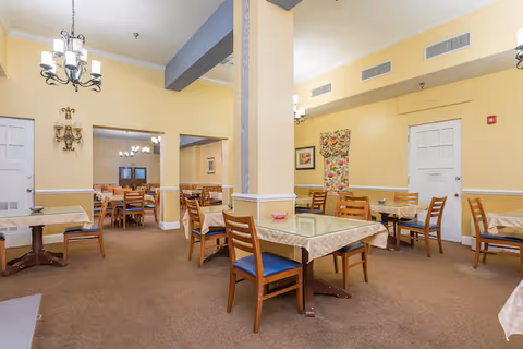 A dining room in Colonial Assisted Living at West Palm Beach featuring multiple wooden tables with beige tablecloths and wooden chairs with blue cushions. The room has yellow walls, carpeted floors, and several light fixtures including chandeliers and wall sconces. There is a floral curtain on one wall and a white door in the background.