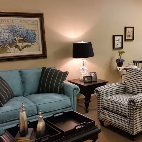 A cozy living room with a light blue sofa adorned with striped pillows, a patterned armchair, a dark wooden coffee table with decorative items, a side table with a clear glass lamp and framed photos, and wall art featuring floral and bird themes.