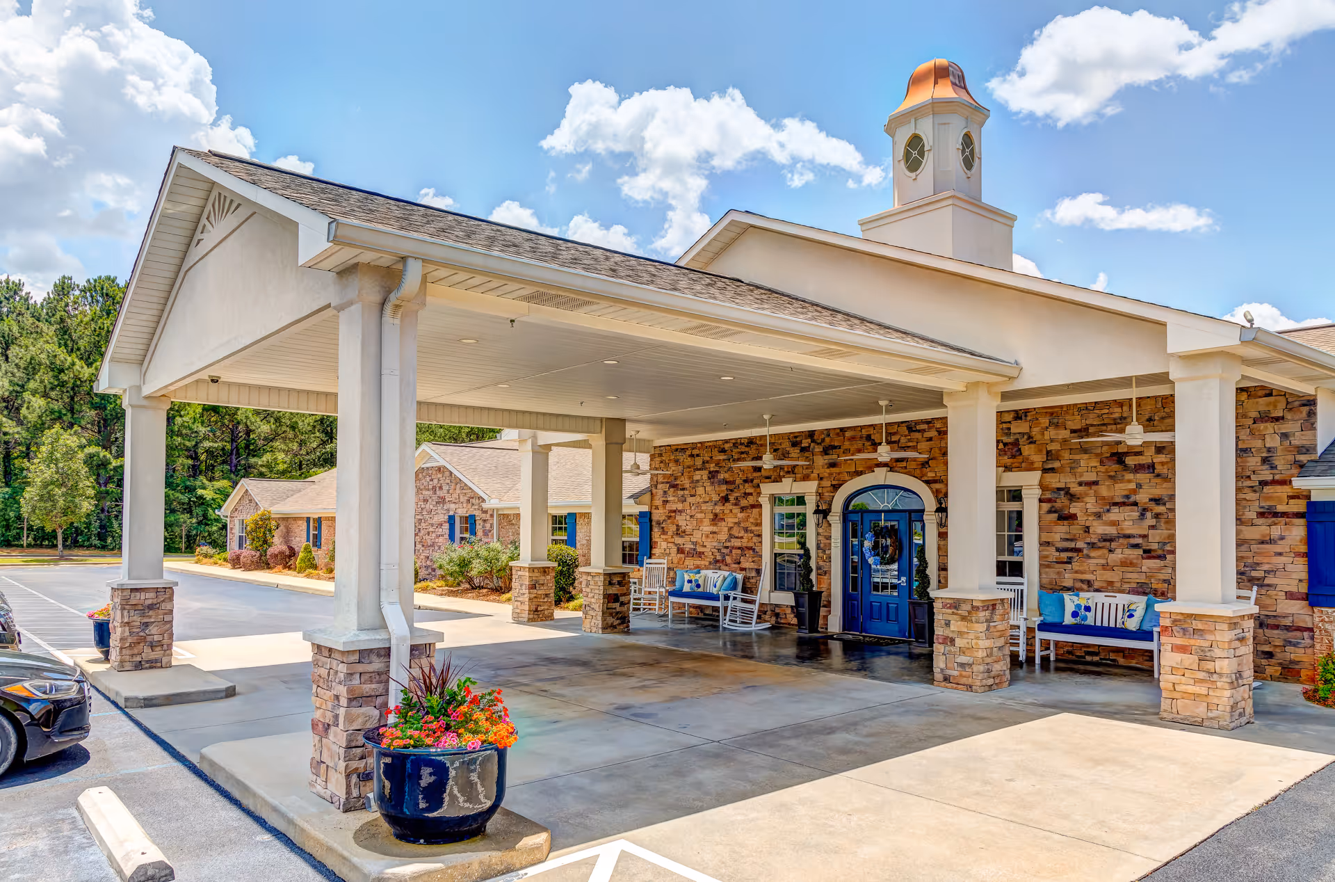Entrance of a senior living facility with a covered drop-off area supported by columns with stone bases. The building has a brick facade, a blue door, benches with cushions, ceiling fans, and a small cupola on the roof. There are flower pots with colorful flowers near the entrance and a parking area adjacent to the building.