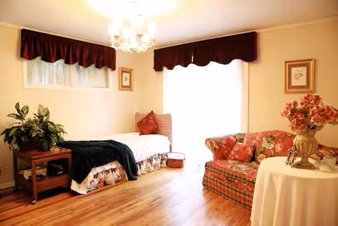 A cozy memory-care bedroom with a bed, floral sofa, plants, and a round table.