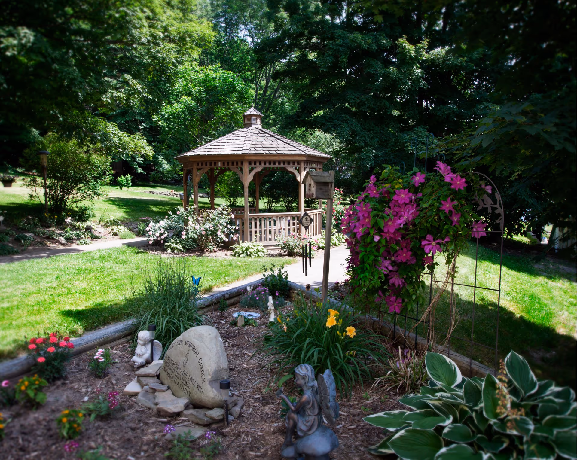 A peaceful garden area with a wooden gazebo surrounded by lush green trees and various flowering plants. There is a stone memorial marker and garden decorations including a small angel statue and wind chimes. The garden is well-maintained with a mix of colorful flowers and greenery.