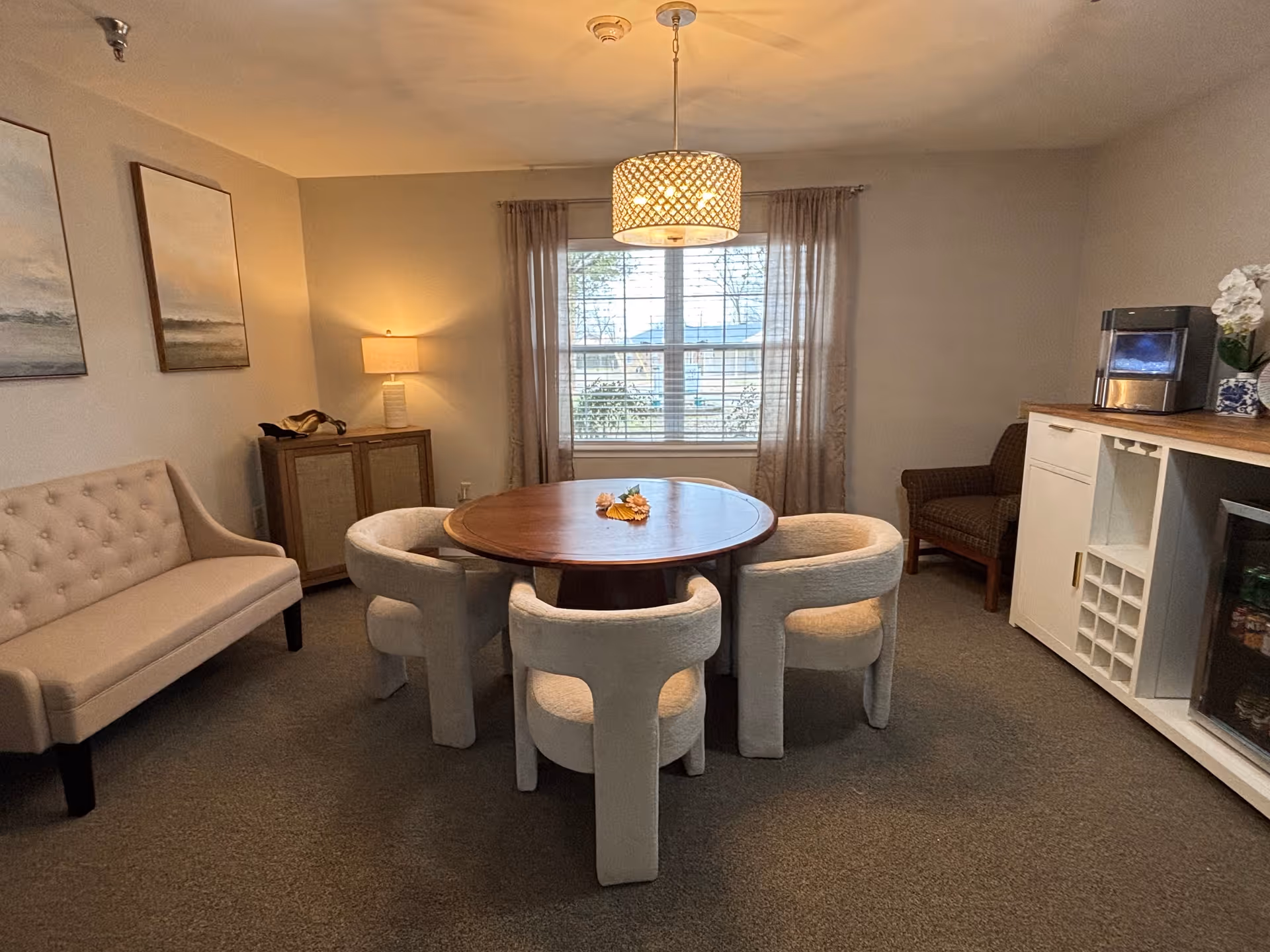A cozy interior room with a round wooden table surrounded by four cushioned chairs. There is a beige tufted bench against the left wall, two framed paintings above it, and a small cabinet with a lamp on top in the corner. A window with sheer curtains lets in natural light. On the right side, there is a white cabinet with a small refrigerator, a wine rack, a coffee machine, and a brown armchair next to it. A decorative hanging light fixture is centered above the table.
