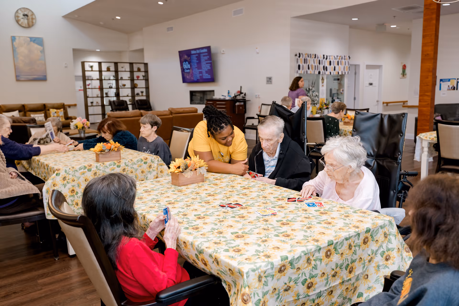 A group of elderly people sitting around tables with sunflower-patterned tablecloths in a common area, playing card games. A caregiver in a yellow shirt is assisting one elderly man. The room has comfortable seating, a wall-mounted TV, and decorative flower arrangements on the tables.