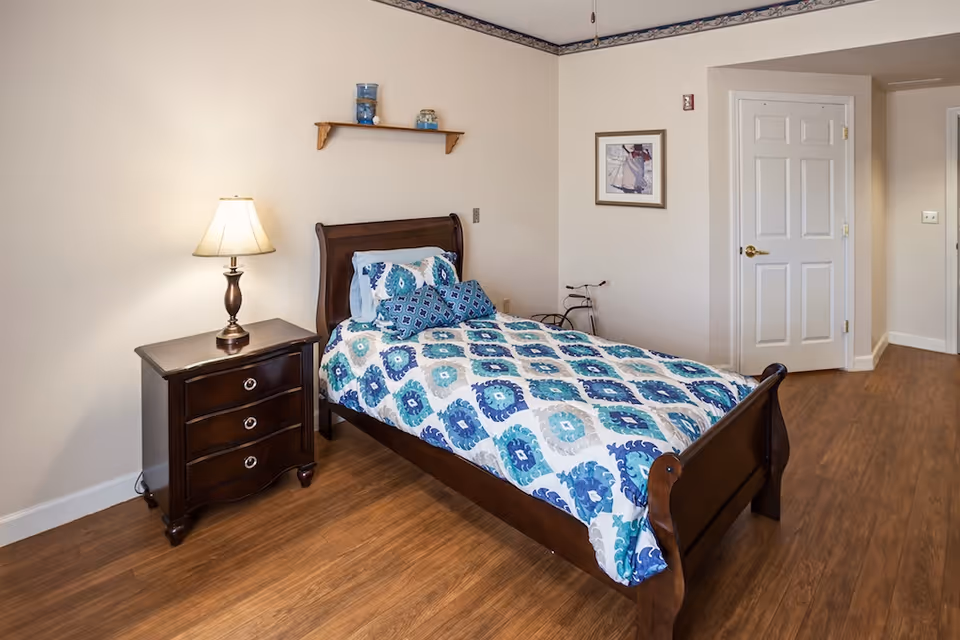 A neatly made single bed with a blue and white patterned comforter and matching pillows in a room with wooden flooring. Next to the bed is a dark wooden nightstand with a lamp on it. A small wooden shelf with decorative items is mounted on the wall above the bed. There is a framed picture on the wall and a closed white door in the background.
