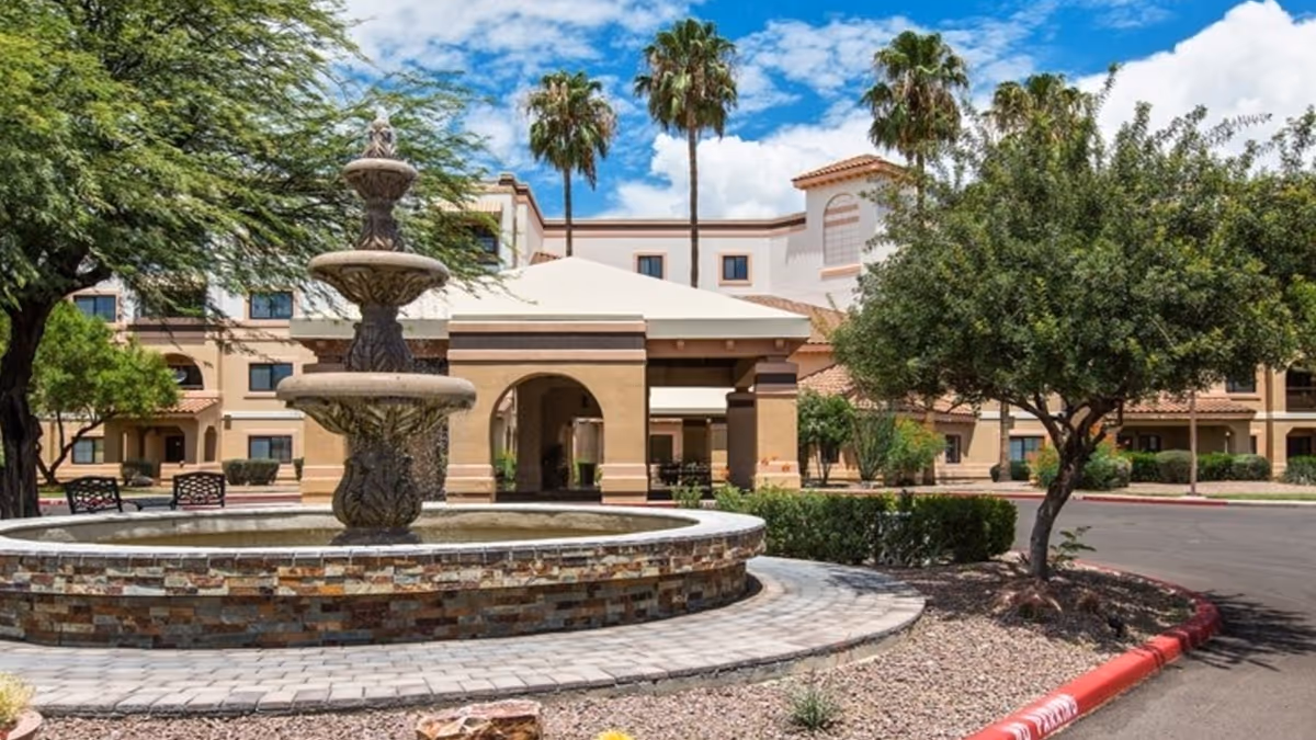 The front entrance of a senior living building with a multi-tiered fountain, covered porte-cochere, palm trees, and a circular driveway.