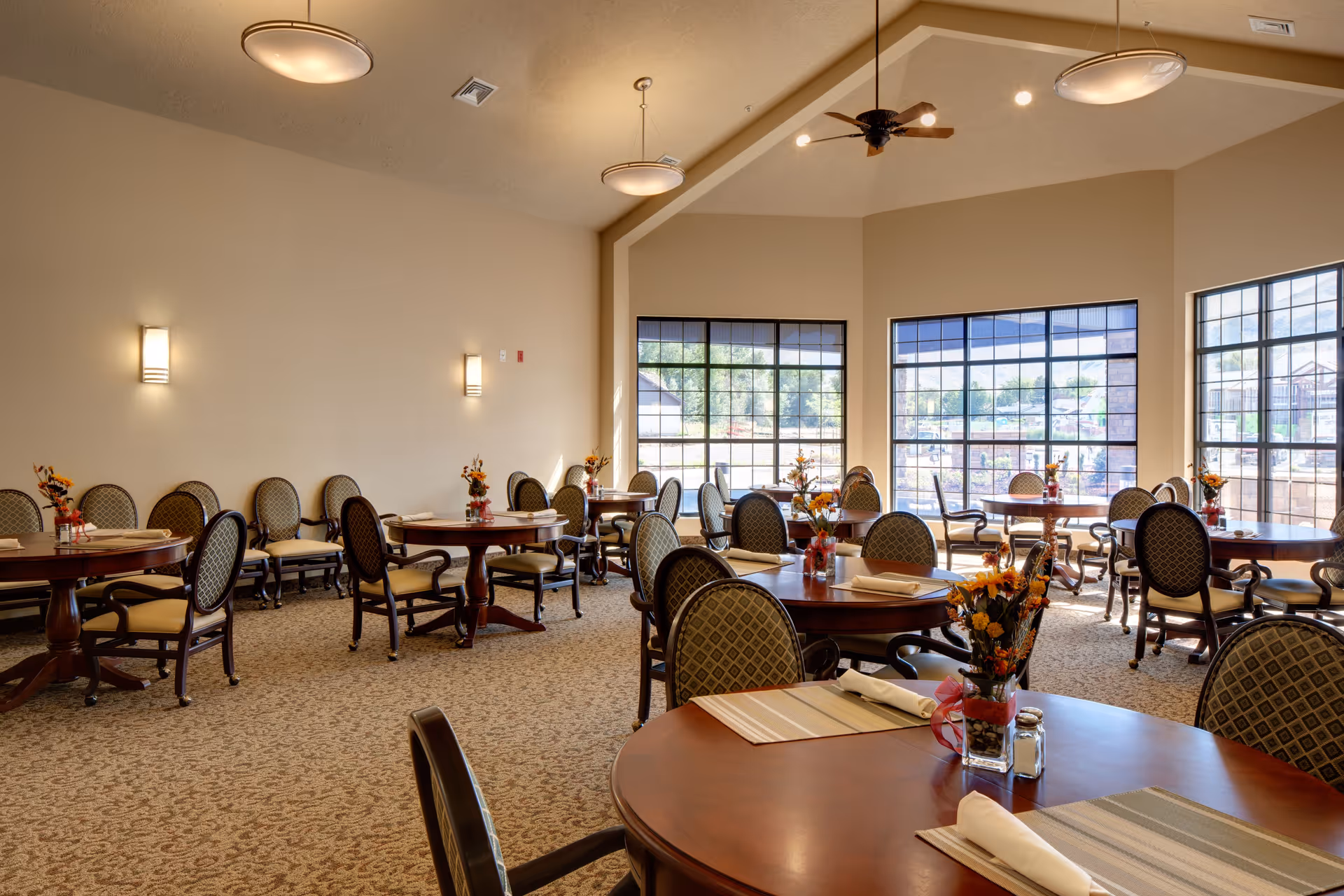 A bright dining room with several round wooden tables and cushioned chairs arranged neatly. Each table has placemats, rolled napkins, and a small floral centerpiece. Large windows allow natural light to fill the room, and ceiling lights and fans provide additional illumination.