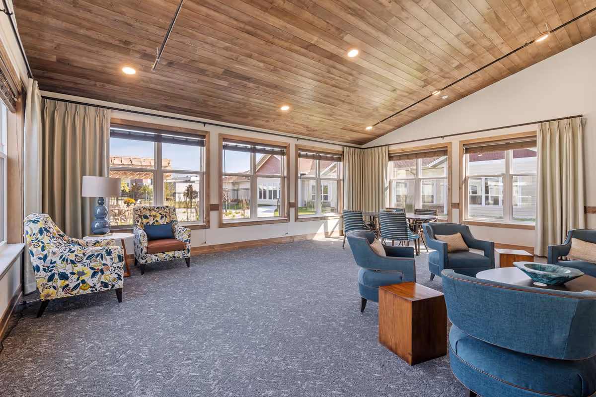 A spacious and well-lit common area with large windows allowing natural light to fill the room. The room features a wooden ceiling with recessed lighting, carpeted floor, and a mix of comfortable seating including floral patterned armchairs, blue upholstered chairs around a circular table, and a small dining table with chairs near the windows. Beige curtains frame the windows, and a blue lamp sits on a side table next to the floral armchairs.