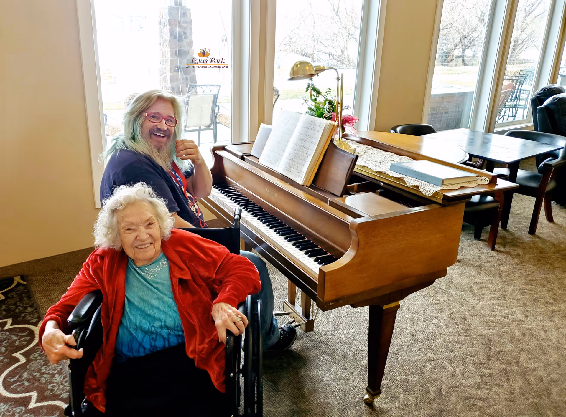 An elderly woman in a wheelchair and a smiling companion sit beside a grand piano in a sunlit senior living common room.