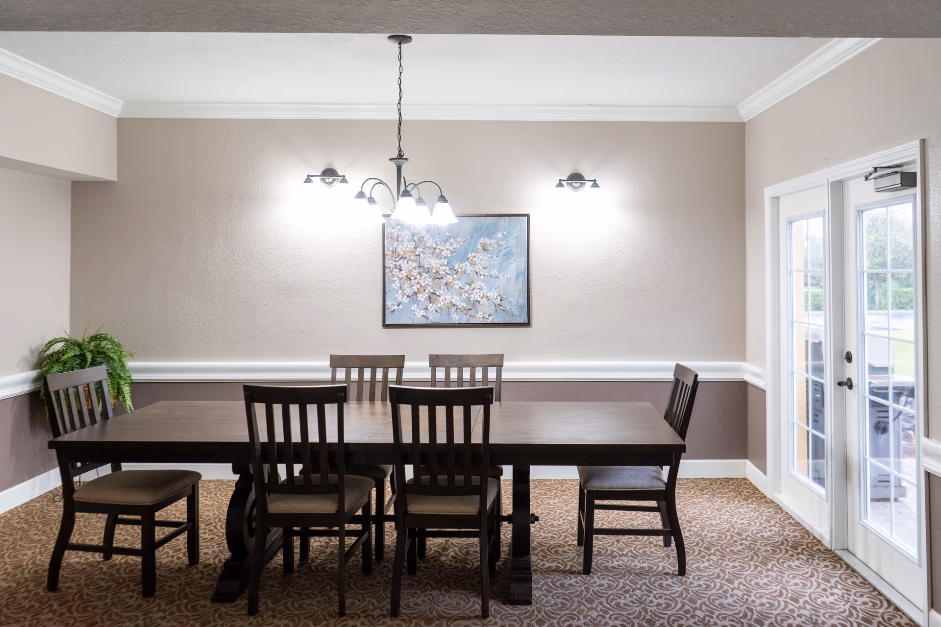A dining room with a dark wooden table and six matching chairs. The walls are painted beige with white trim, and there is a painting of white flowers on a blue background hanging on the wall. Above the table, a chandelier with five lights is hanging, and two wall sconces provide additional lighting. To the right, there are glass double doors leading outside. A green potted plant is placed in the corner on the left side.