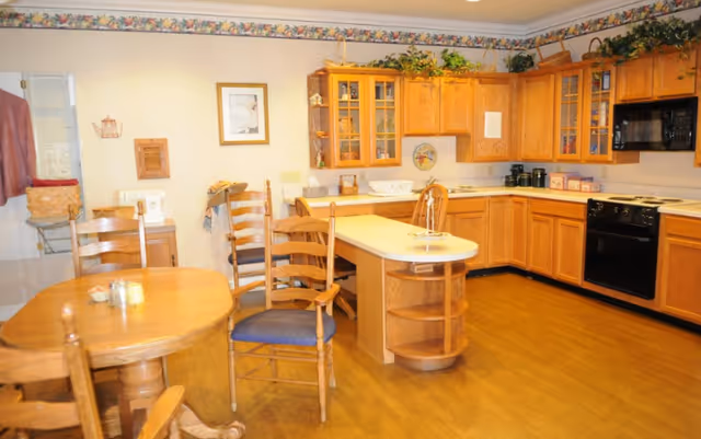 A spacious kitchen and dining area with wooden cabinets, a kitchen island, and a round wooden dining table with chairs. The kitchen features black appliances including a microwave and oven. There are decorative plants on top of the cabinets and framed artwork on the wall.