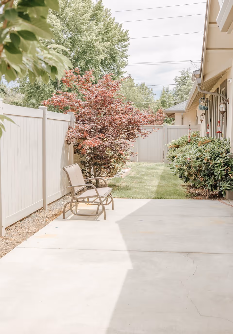Outdoor patio area with a metal bench, surrounded by a white fence, green bushes, and a tree with red leaves. The patio is adjacent to a building with windows and hanging lanterns.