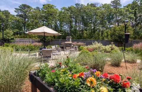 Outdoor garden area with colorful flowers in the foreground, patio seating with tables and chairs under a large umbrella, a stone fireplace, and a backdrop of tall trees and a wooden fence.