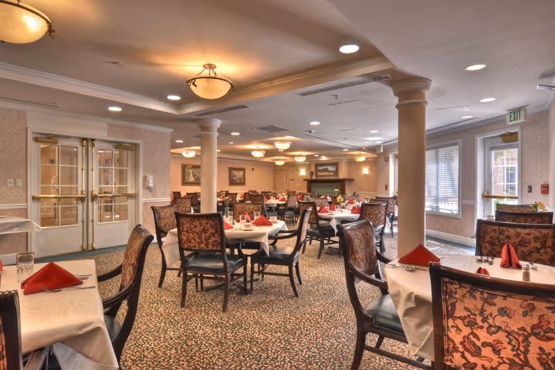 A dining room in a senior living facility with multiple tables covered in white tablecloths, each set with red folded napkins, glasses, and silverware. The room features patterned carpet, upholstered chairs, columns, ceiling lights, and large windows letting in natural light.