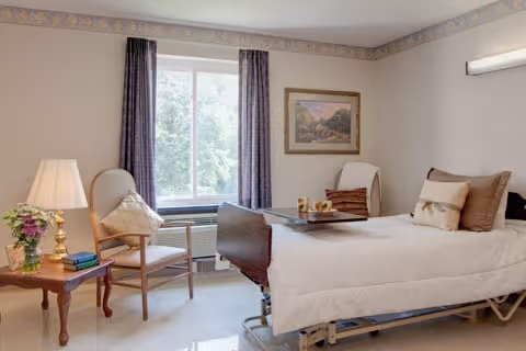 A bright and clean patient room with a hospital bed, a wooden side table with a lamp and flowers, two chairs near a window with purple curtains, and a framed picture on the wall.