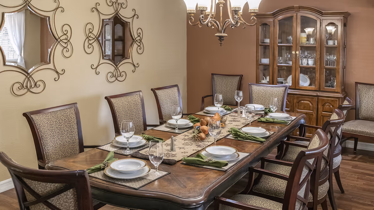 A formal dining room with a wooden dining table set for eight people. The table is decorated with a beige table runner, green cloth napkins, white plates, and clear wine glasses. The room features upholstered chairs with wooden frames, a wooden china cabinet filled with dishes and glassware, two decorative mirrors on the wall, and a chandelier hanging above the table.