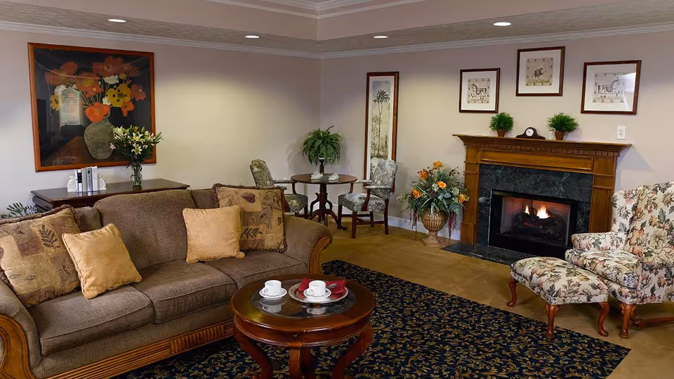 A cozy living room with a brown sofa adorned with patterned and solid cushions, a round wooden coffee table with two white teacups and saucers, a floral upholstered armchair with matching ottoman, a fireplace with a wooden mantel and a fire burning inside, framed artwork and plants decorating the walls and corners, and a small round table with two chairs in the background.