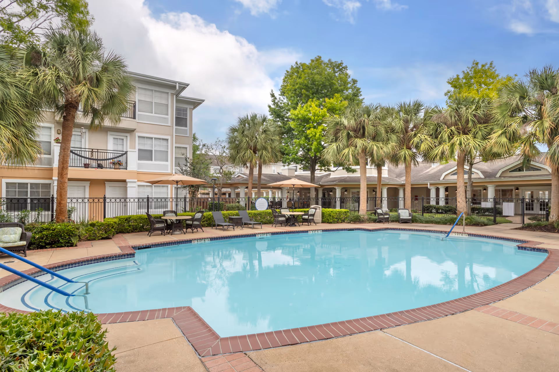 Outdoor swimming pool surrounded by lounge chairs, umbrellas, palm trees and a multi-story residential building.
