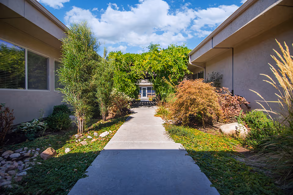 A concrete walkway between two beige buildings with landscaped greenery on both sides, including bushes, small trees, and rocks. At the end of the walkway, there is a green leafy archway with a door visible beyond it. The sky is blue with scattered clouds.