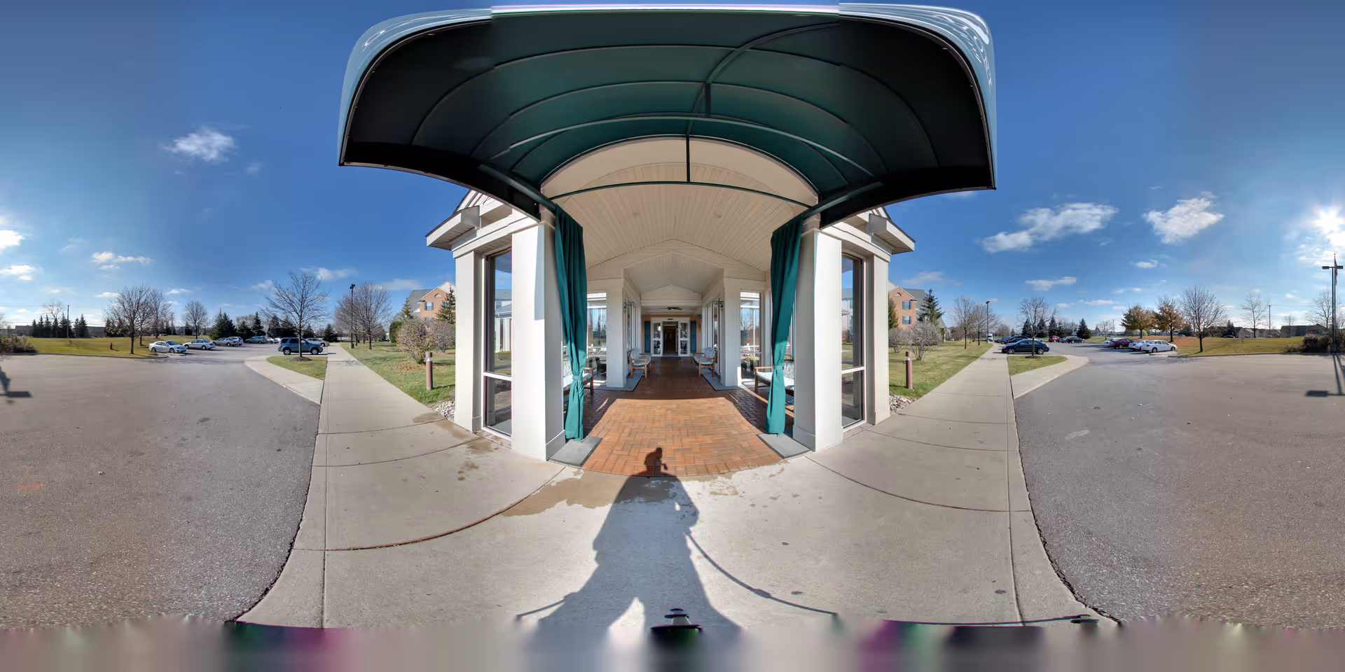 Entrance to Oakmont Sterling Senior Community with a covered walkway supported by white pillars and green curtains, leading to glass doors. The surrounding area includes a parking lot with several cars and grassy areas with trees under a clear blue sky.