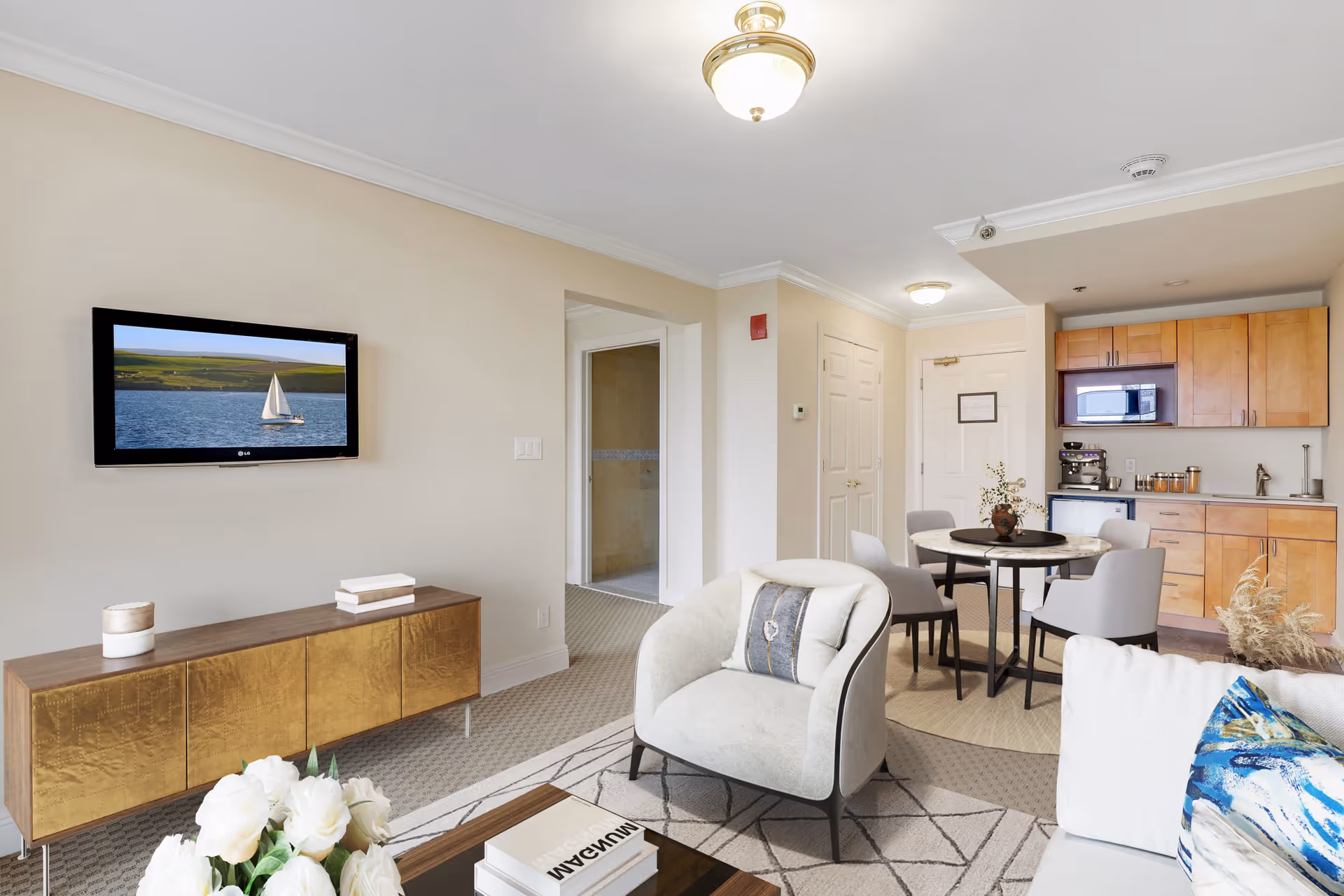 A bright and modern living area in a senior living facility featuring a wall-mounted flat-screen TV displaying a sailboat on water, a wooden cabinet below the TV, a comfortable armchair with a decorative pillow, a round dining table with four chairs, and a kitchenette with wooden cabinets, a microwave, and a coffee machine. The room has beige walls, carpeted flooring, and ceiling lights.