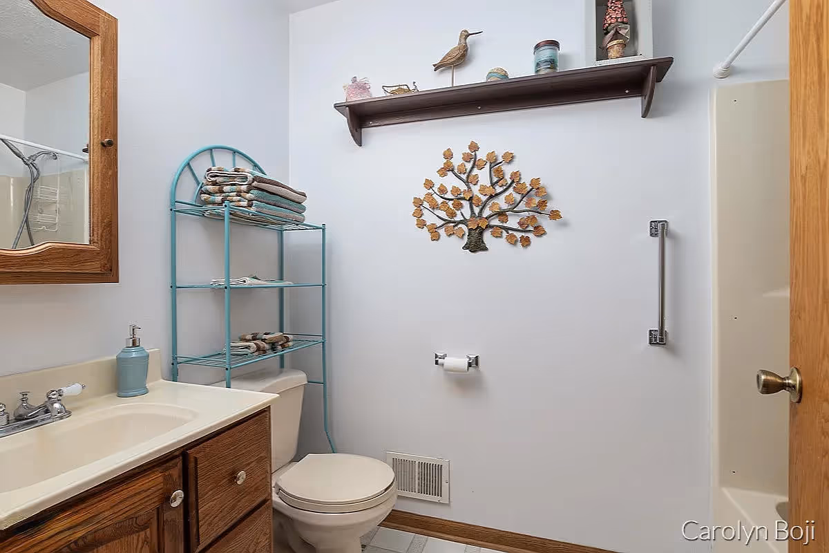 A small bathroom with a wooden vanity and sink on the left, a toilet with a teal metal shelf above it holding folded towels, a wall-mounted decorative metal tree, a wooden shelf with decorative items above it, and a shower with a grab bar on the right.