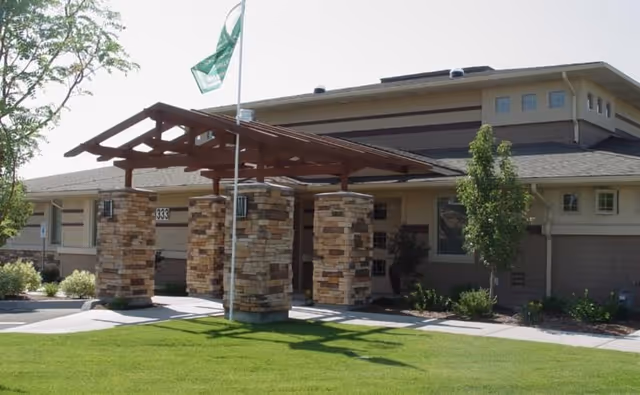Exterior view of a single-story assisted living facility building with a covered entrance supported by stone pillars, a flagpole with a green flag, and well-maintained green lawn and landscaping.