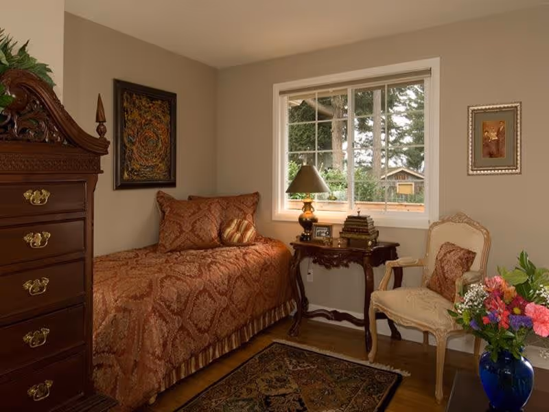 Cozy single bedroom with an ornate wooden dresser, patterned bedspread, side table with a lamp, and a chair by the window.