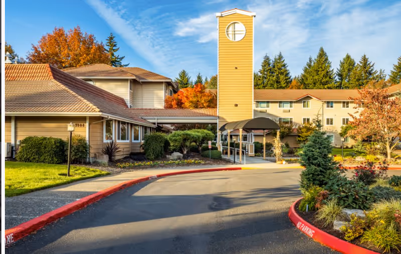 Exterior view of Van Mall Retirement Vancouver facility showing a multi-story building with beige siding and a prominent yellow clock tower. The entrance is covered with a canopy, surrounded by landscaped greenery, trees with autumn foliage, and a curved driveway with red curbs.