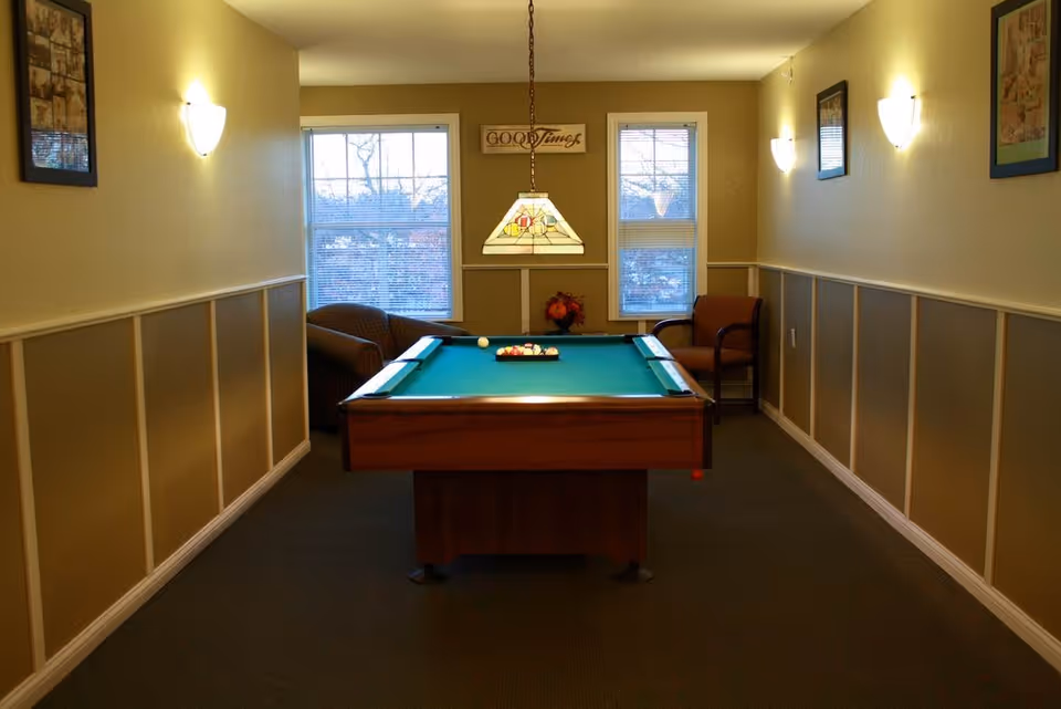 A cozy game room with a pool table centered in the room. The pool table has a green felt surface with billiard balls arranged for a game. There are two windows at the far end letting in natural light, with a decorative hanging lamp above the pool table. Two chairs are placed near the windows, and the walls are painted beige with white wainscoting and framed pictures. Wall sconces provide additional lighting.