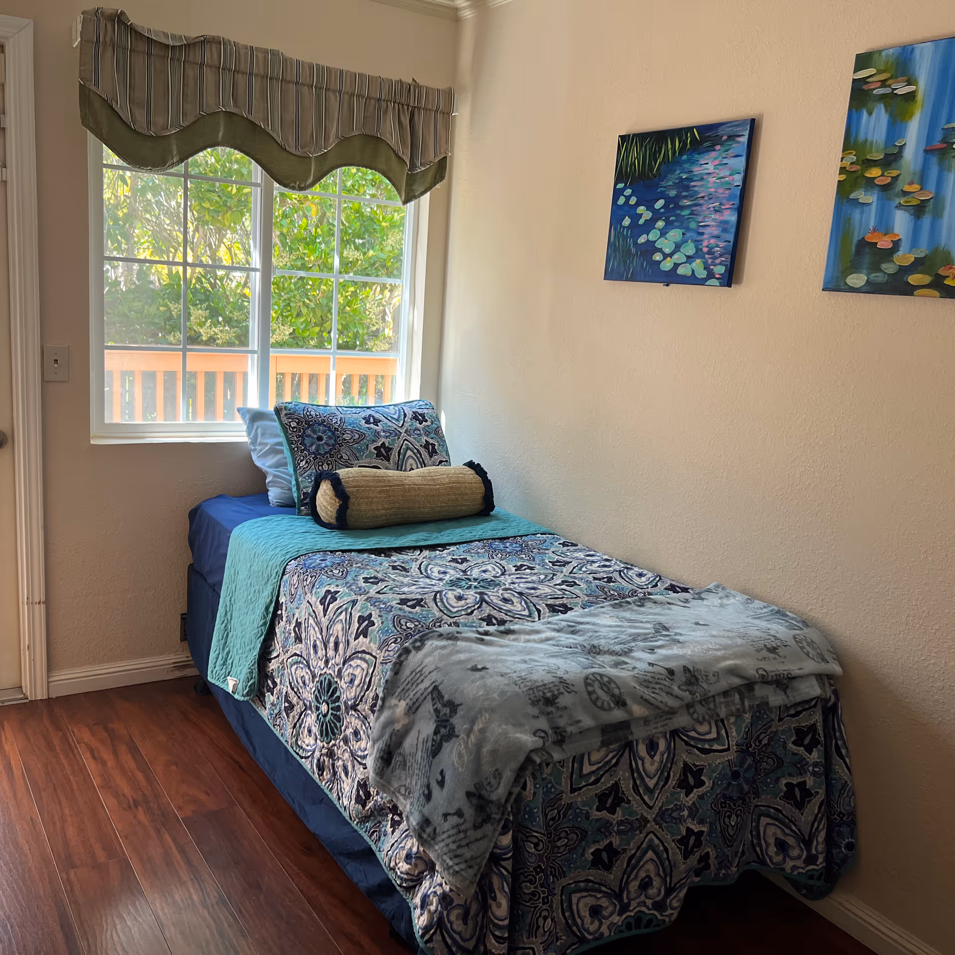 A tidy single bedroom with patterned bedding by a window and two paintings on the wall.