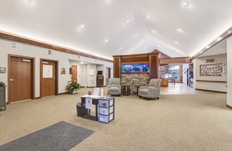Bright, spacious senior living lobby with a vaulted ceiling, two armchairs by a central aquarium, and doors along the back wall.
