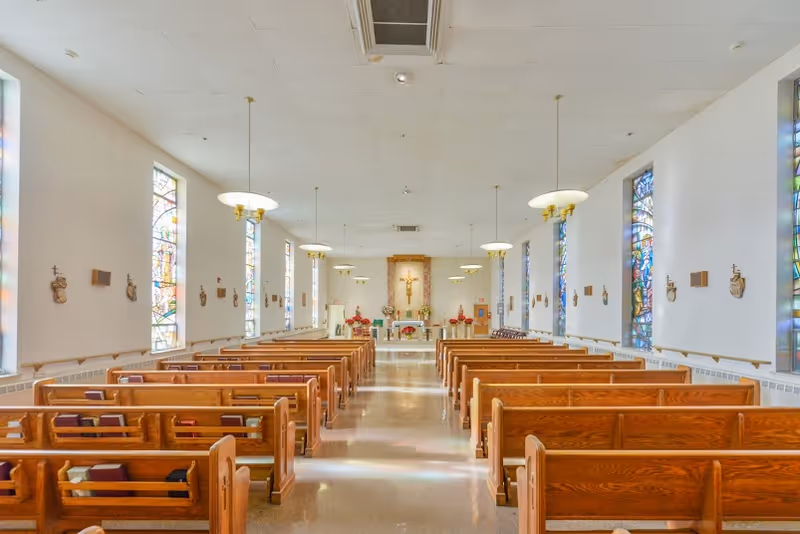 Interior view of a chapel with wooden pews arranged in rows facing an altar with a crucifix. The chapel has tall stained glass windows on both sides and hanging light fixtures from the ceiling.