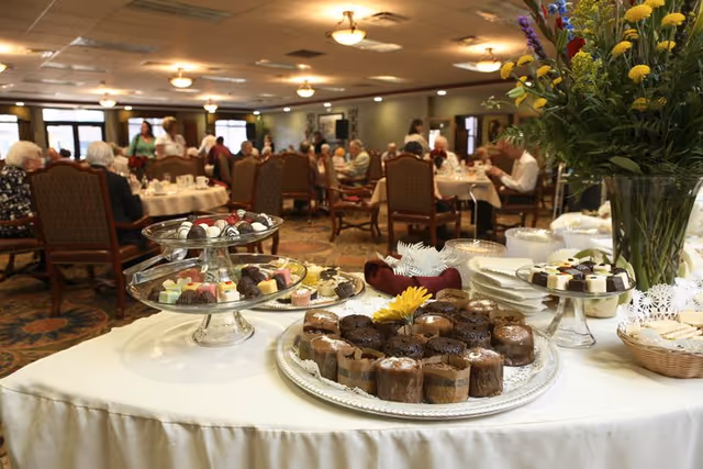 A dessert table with various cakes, chocolates, and cookies in the foreground, with elderly people seated at round dining tables in the background inside a well-lit dining room.
