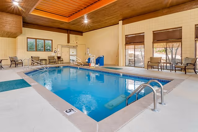 Indoor swimming pool with clear blue water surrounded by a beige tiled deck. Several chairs and small tables are placed along the walls under large windows with brown blinds. The ceiling is wooden with recessed lighting, and there is a pool lift for accessibility.