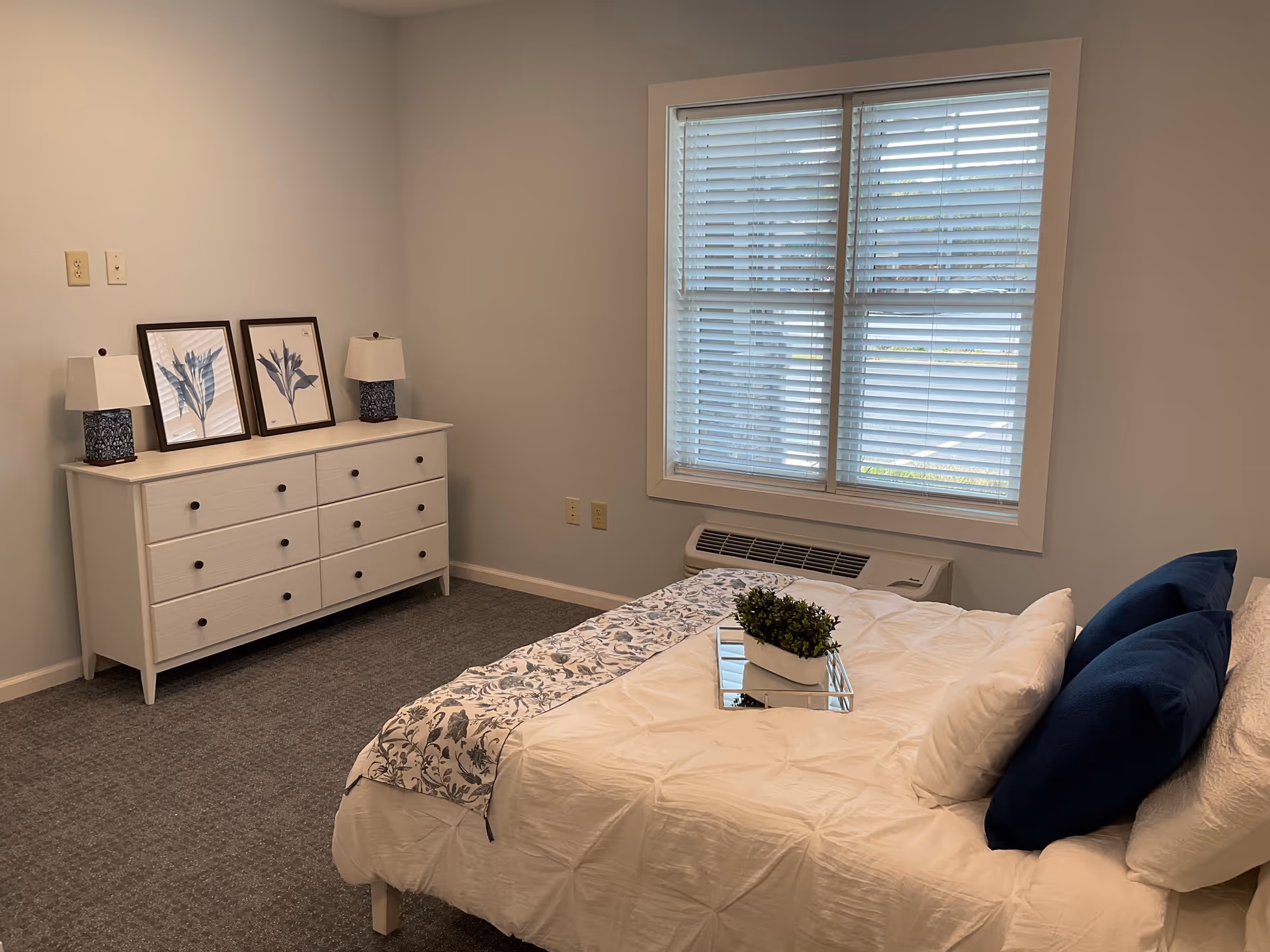A bedroom with a white bed featuring blue and white pillows and a floral bed runner. A small plant on a tray is placed on the bed. There is a white dresser with six drawers against the wall, decorated with two framed botanical prints and two table lamps with blue bases and white shades. A window with white blinds is above a heating/cooling unit on the wall.
