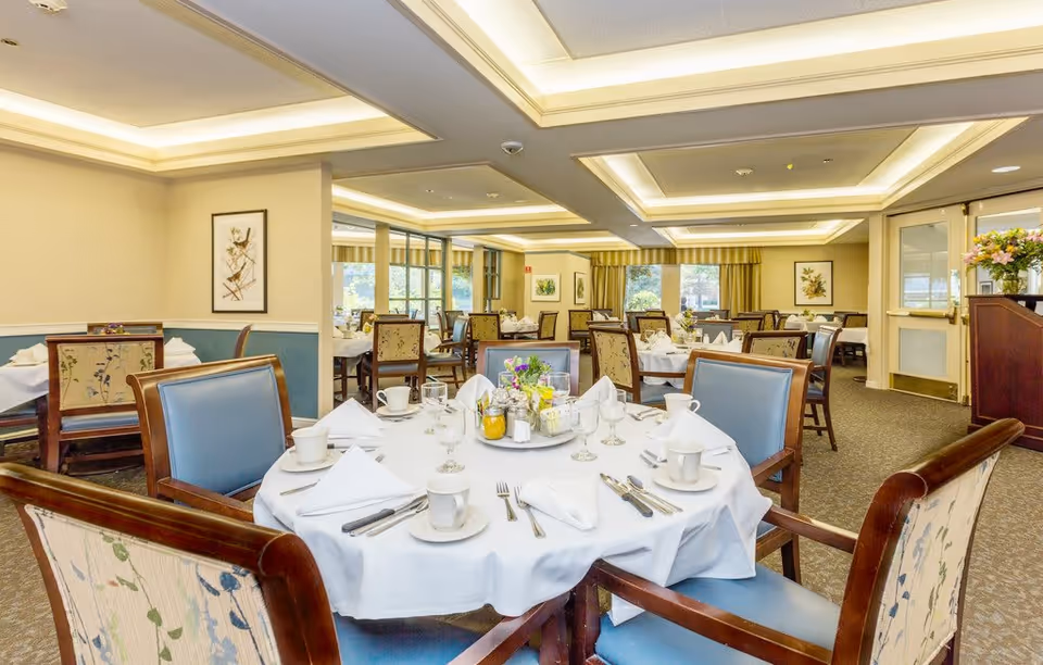 A well-lit dining room with round tables covered in white tablecloths, set with cups, glasses, silverware, and napkins. The chairs have wooden frames with blue cushions and floral-patterned backs. The room has beige walls, framed artwork, large windows with curtains, and a carpeted floor.