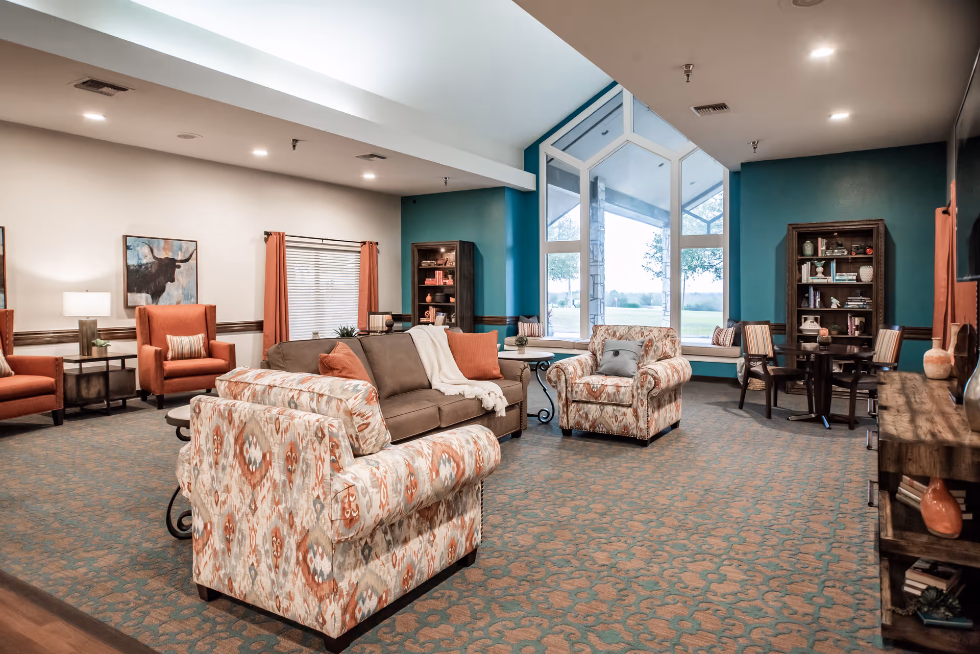 A spacious and well-lit living room area in a senior living facility with patterned armchairs, a brown sofa with orange and white pillows, a large window with a view of the outdoors, bookshelves, a small round table with chairs, and warm lighting.