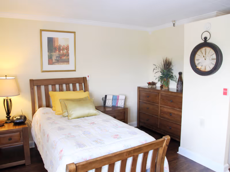 A cozy bedroom with a wooden bed frame and a floral bedspread. There are two pillows on the bed, one yellow and one green. To the left of the bed is a wooden nightstand with a lamp and a clock radio. On the right side of the room is a wooden dresser with decorative items and a large round wall clock above it. The walls are light-colored and there is a framed picture hanging above the bed.