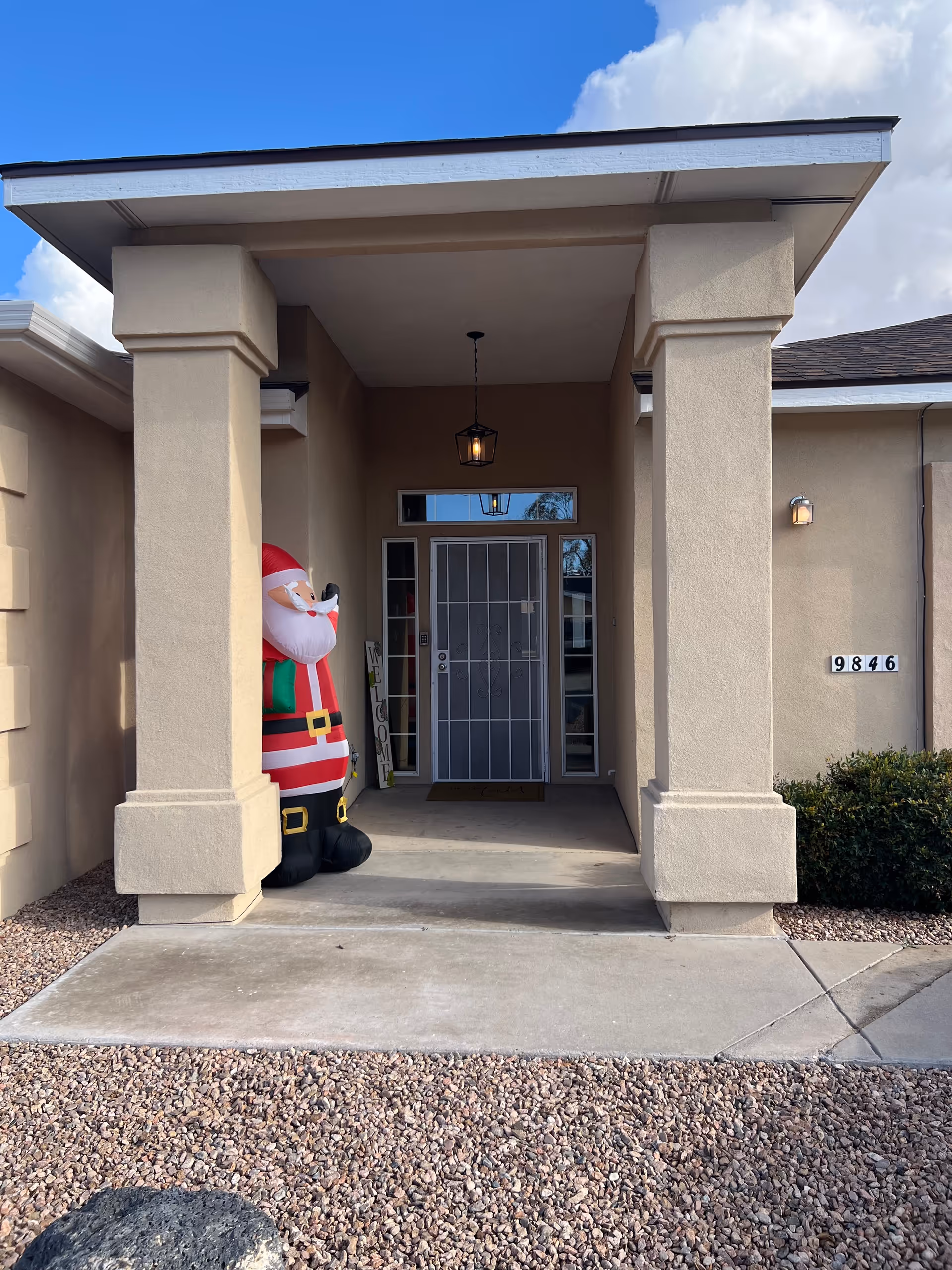 Entrance of a beige building with two large square columns supporting a covered porch. A large inflatable Santa Claus decoration is placed on the left side near the door. The door has a metal security screen and a hanging lantern light above it. The building number 9846 is visible on the right wall next to some bushes. The ground is covered with small rocks and a concrete walkway leads to the door.