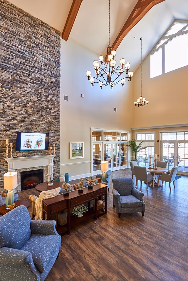 Spacious high-ceiling common living area with a stone fireplace, chandeliers, upholstered chairs and a dining table.