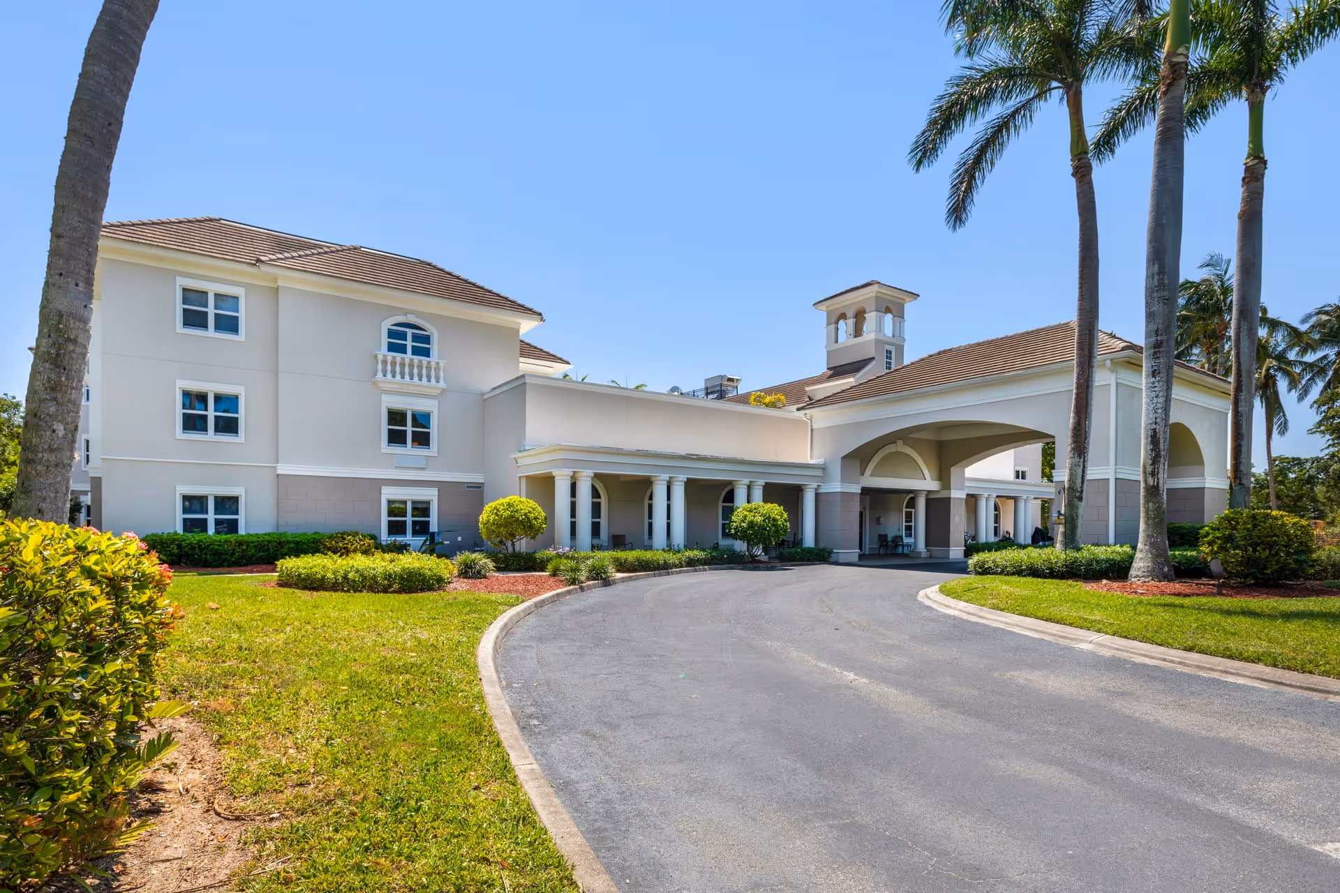 Exterior view of a senior living facility building with a covered entrance, surrounded by palm trees and landscaped greenery under a clear blue sky.